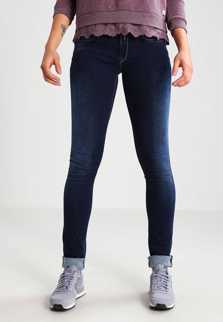 Jeans ajustados de color azul oscuro hechos de denim, con una cintura alta y un corte hasta el tobillo, combinados con zapatillas gris claro y un suéter morado.