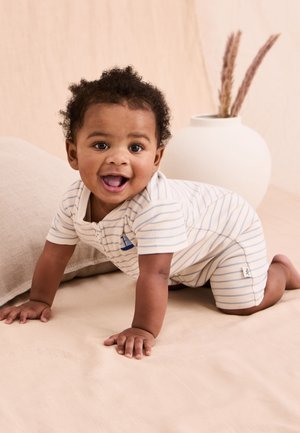 Smilende baby kravler på beige overflade iført hvid og blåstribet romper med pude i neutrale toner og hvid vase med tørrede planter i baggrunden.