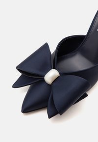 Chaussure bleu marine à bout pointu avec un grand nœud en tissu et un détail en perle blanche sur un fond clair uni.