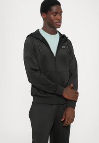 Svart zip-up hoodie med en mjuk textur, med en huva med dragsko, dragkedja framtill och EA7-logotyp på bröstet. Åtföljs av matchande byxor.