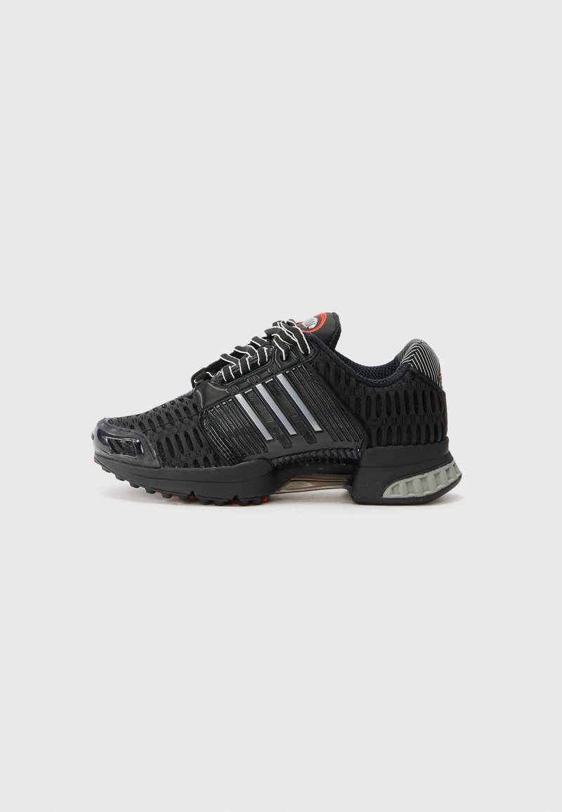 Chaussure de sport noire avec une tige en mesh, des bandes réfléchissantes, une semelle en caoutchouc et des accents rouges. Design distinctif respirant et tissu texturé.