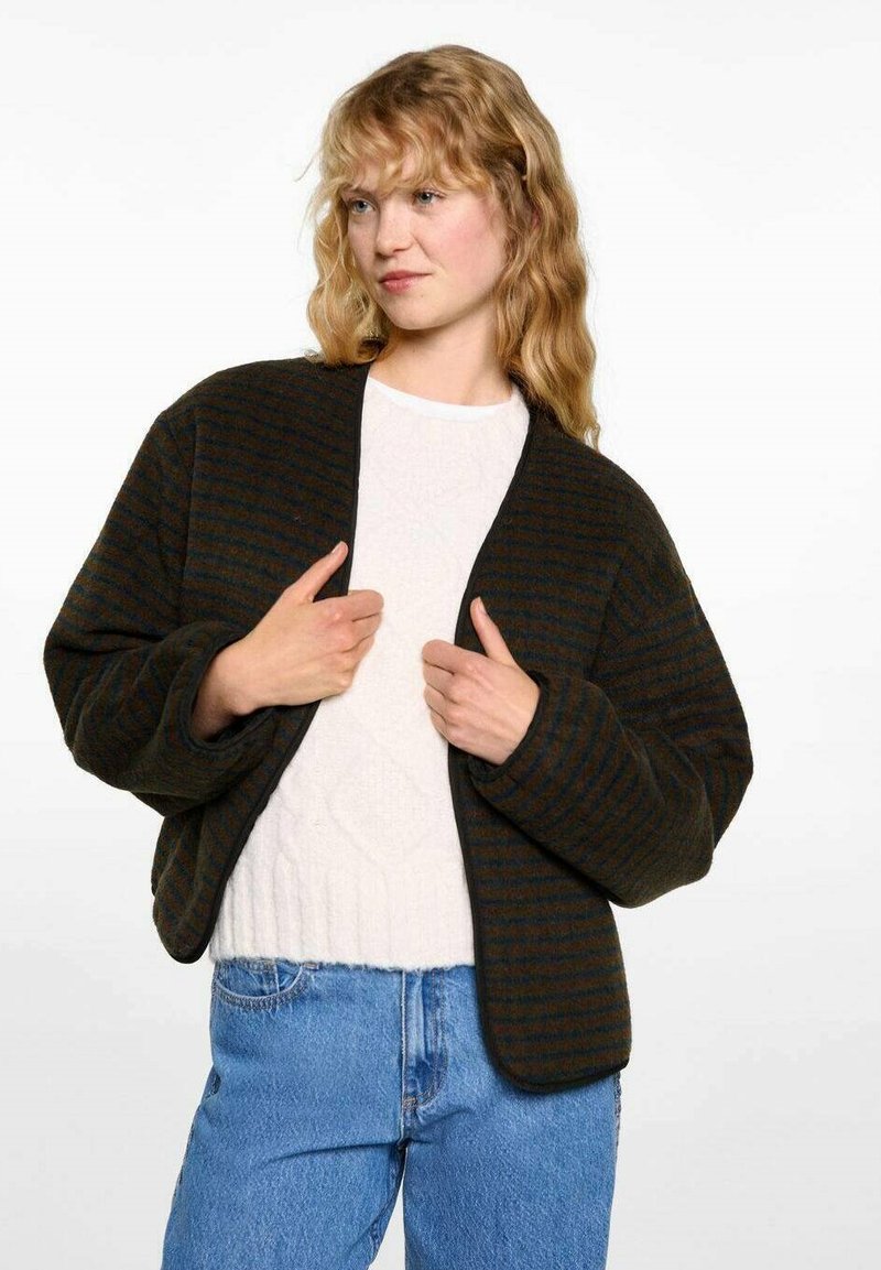 Kaki- och mörkbrun randig oversized jacka med en avslappnad passform och vida ärmar, bärs över en ljus tröja, tillsammans med blåjeans.