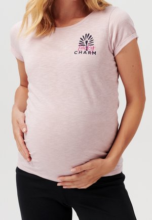 Gravid kvinde, der holder om sin mave, iført en lyserød T-shirt med teksten "Turkey Charm" og en stiliseret kalkun grafik på brystet.