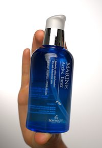 Blå glassflaske med pumpehette, merket "Marine Active Toner." Teksten beskriver fuktighetsgivende og hydrerende egenskaper. Glatt overflate.