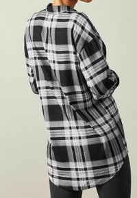 Chemise à carreaux noir et blanc à manches longues, avec un ourlet arrondi et une coupe décontractée, mettant en avant un mélange de rayures verticales et horizontales.