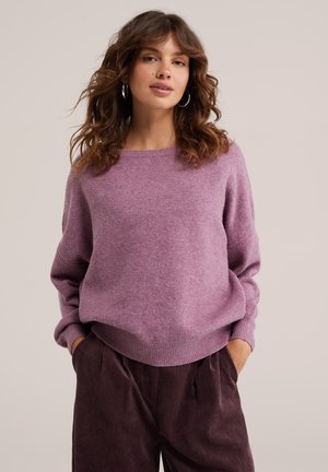 Woman with curly hair wears a purple sweater and dark brown corduroy pants, standing with hands in pockets against a plain background.