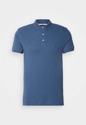 Blue short-sleeve polo shirt, made of cotton, with a three-button placket and a small logo patch on the chest. Smooth texture, classic design.