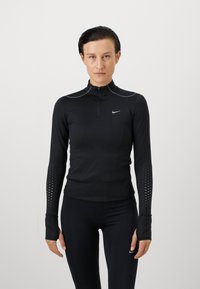Czarna koszulka sportowa z długim rękawem i kołnierzem na zamek błyskawiczny do połowy, z odblaskowymi akcentami i teksturowanymi kropeczkami na rękawach. Wykonana z elastycznego materiału.