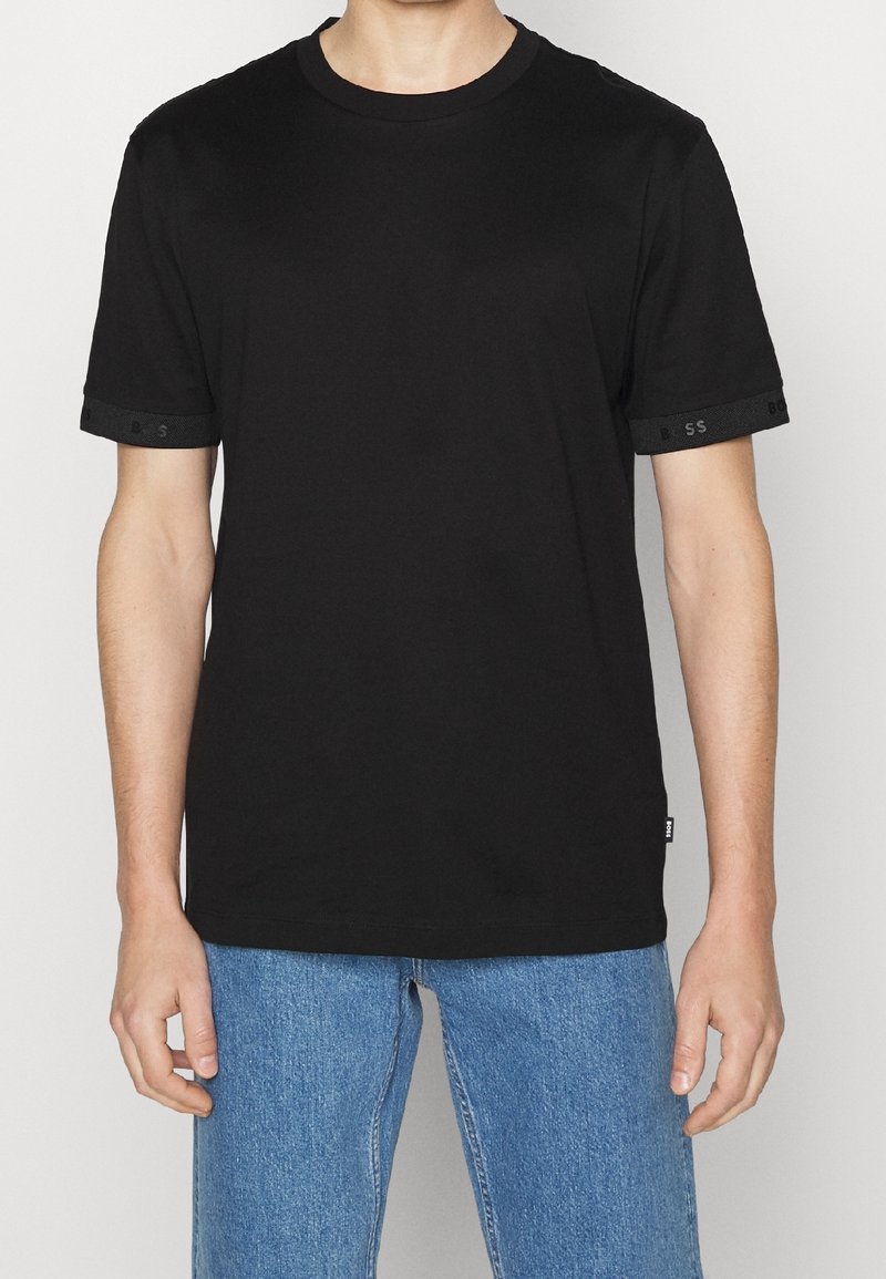 T-shirt en coton noir avec un col rond, des manches courtes agrémentées de bordures texturées noires et un petit logo sur la manche.