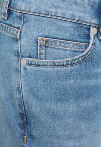 Světle modré džíny z denimu s hladkým povrchem, obsahující malou přední kapsu a hardware se stříbrnými knoflíky. Kontrastní oranžové prošívání.