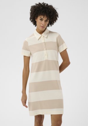 Ženska s kodrastimi lasmi, oblečena v poletno polo obleko z bež in belimi vodoravnimi črtami z kratkimi rokavi, stoji pred enobarvnim ozadjem.