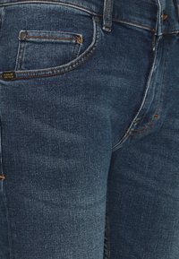 Jeans i mörkblå denim med en klassisk femfickorsdesign, kontrasterande orange sömmar och en mjuk, något texturerad yta.