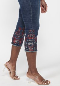 Dark denim cropped pants with floral embroidery in red and white at the hem. Finished with a frayed cuff. Paired with clear heeled sandals.
