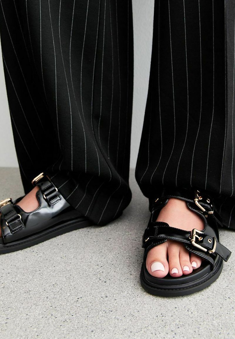 Svarta sandaler med glänsande material, dubbla spännen och öppen tå-design, tillsammans med randiga vida byxor.