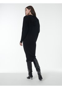 Schwarzes, enganliegendes Strickkleid mit Kapuze, das über eine gerippte Textur und lange Ärmel verfügt. Kombiniert mit kniehohen schwarzen Stiefeln mit Absatz.