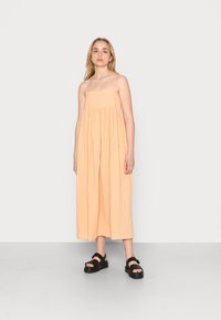Vestido maxi sem mangas, na cor pêssego, com alças finas e um design solto e franzido, combinado com sandálias de plataforma pretas.