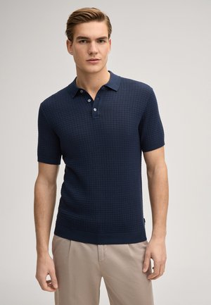 Joven con polo de manga corta azul marino texturizado y pantalones beige, sobre un fondo claro y liso.