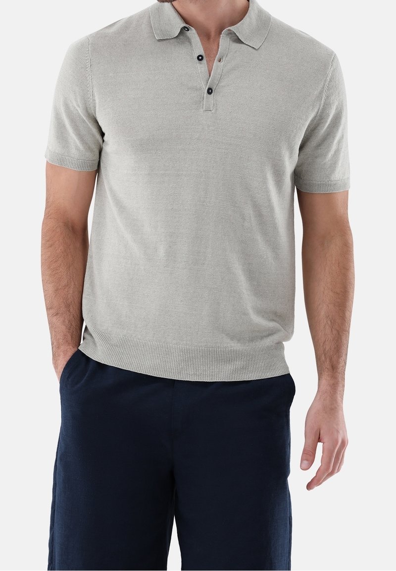 Mężczyzna ubrany w jasnoszary polo z krótkim rękawem z guzikami i ciemnoniebieskie spodnie, stojący z jedną ręką w kieszeni na tle białym.