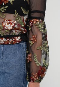 Top trasparente nero con ricami floreali in rosso, verde e giallo. Presenta maniche lunghe a sbuffo e un dettaglio a righe ai polsini.