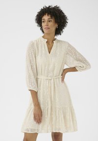 Un vestido de color crema con escote en V, mangas tres cuartos y un cinturón de lazo. Cuenta con una falda de capas y un patrón floral texturizado en toda su superficie.