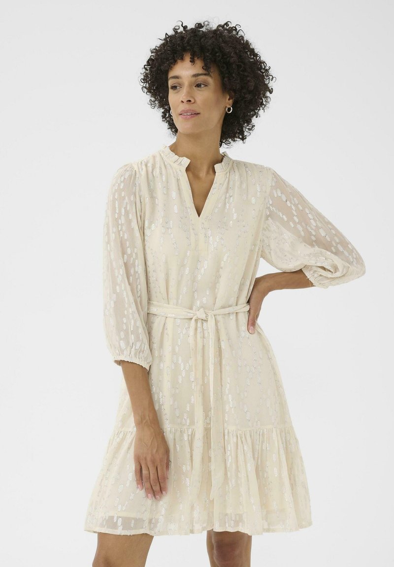 Un vestido de color crema con escote en V, mangas tres cuartos y un cinturón de lazo. Cuenta con una falda de capas y un patrón floral texturizado en toda su superficie.
