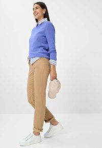 Hellblauer Strickpullover über einem weiß-blauen gestreiften Hemd, kombiniert mit beigen Chinos und weißen Turnschuhen. Hält eine hellbeige Mütze.