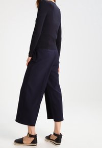 Kvinna med marinblå vida cropped-byxor, svart långärmad topp och svarta espadrille-sandaler med ankelsnöre, stående mot en vit bakgrund.