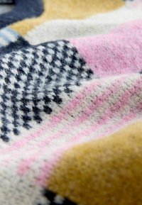 Zbliżenie na miękką tkaninę wełnianą z kolorowymi wzorami, w tym granatową pepitkę, różowe paski oraz fragmenty w kolorze musztardowej żółci.