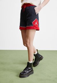 Bermudas deportivas negras y rojas con cintura ajustable mediante cordón, con un contorno blanco y un detalle de logo, combinadas con botas negras gruesas con cordones.