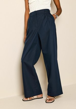 Pantaloni a palazzo blu navy realizzati in un tessuto morbido, con vita alta, pince e tasche laterali. Indossati con sandali beige con cinturino.