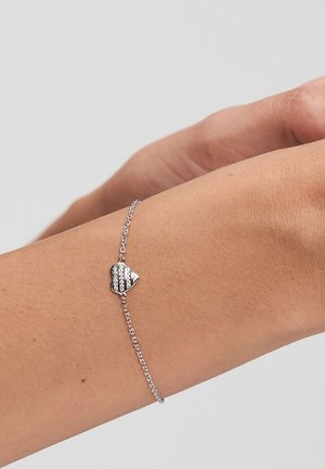 Zierliches Silberarmband mit einem kleinen herzförmigen Anhänger, der mit klaren Kristallen verziert ist, getragen an einem Handgelenk vor einfachem Hintergrund.