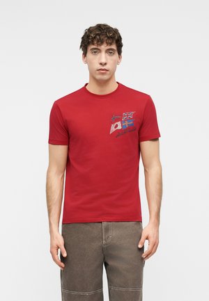 Jonge man met krullend haar die een rood t-shirt draagt met Noordse en Japanse vlaggen op de borst en een bruine broek.