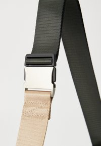 Fibbia in metallo a rilascio laterale che collega cinghie intrecciate nere e beige su uno sfondo neutro.
