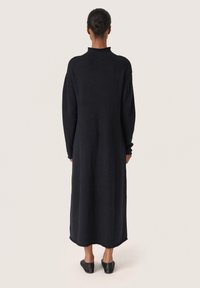 Rochie lungă neagră cu gât înalt și mâneci lungi, fabricată dintr-un material moale. Tivul este ușor curbat. Asortată cu pantofi negri slip-on.