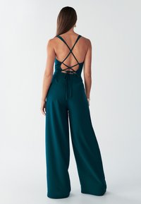 Teal jumpsuit, jossa on ristikkäinen selkäosa, nauhakiristys, leveät lahkeet ja sileä pinta. Ei näkyviä kuvioita tai metalliosia.