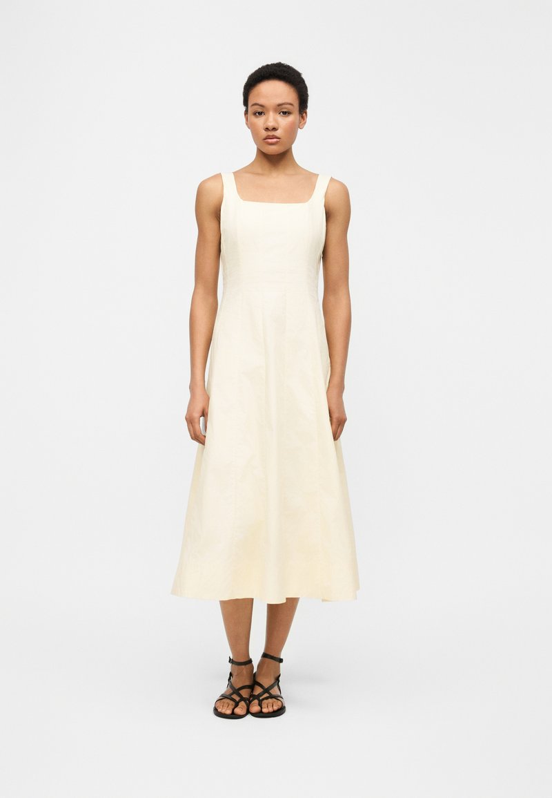 Sieviete stāv ar seju pret kameru, tērpusies krēmkrāsas, bez piedurknēm, kvadrātveida izgriezuma midi kleitā un melnās, atvērtās, plakanās sandales ar siksniņām uz vienkārša fona.