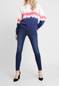 Femme portant un sweat-shirt à manches longues tie-dye aux couleurs rouge et bleu, un jean skinny bleu foncé et des chaussures à talons noirs, debout avec la main sur la hanche.
