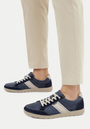 Tmavě modré kožené tenisky s světle šedými detaily, plochými tkaničkami a texturovanou gumovou podrážkou, spárované se světle béžovými krátkými kalhotami.