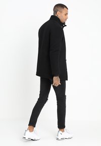 Schwarzer Wollmischungs-Mantel mit hohem Kragen, vorderen Knöpfen und zwei Seiten Taschen, kombiniert mit schwarzen zerrissenen Jeans und weißen Sneakern.