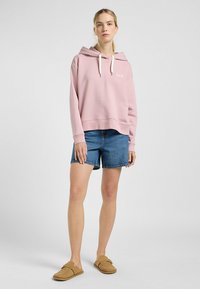 Roze hoodie met een trekkoord en een logo aan de voorkant, gecombineerd met blauwe denim shorts en tan schuifsandalen. Soepele textuur, casual pasvorm.