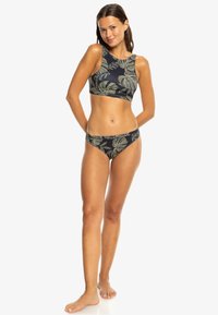 Žena v čiernom dvojdielnom plavkách s zelenými tropickými listami, stojí bosá s rukami za chrbtom.