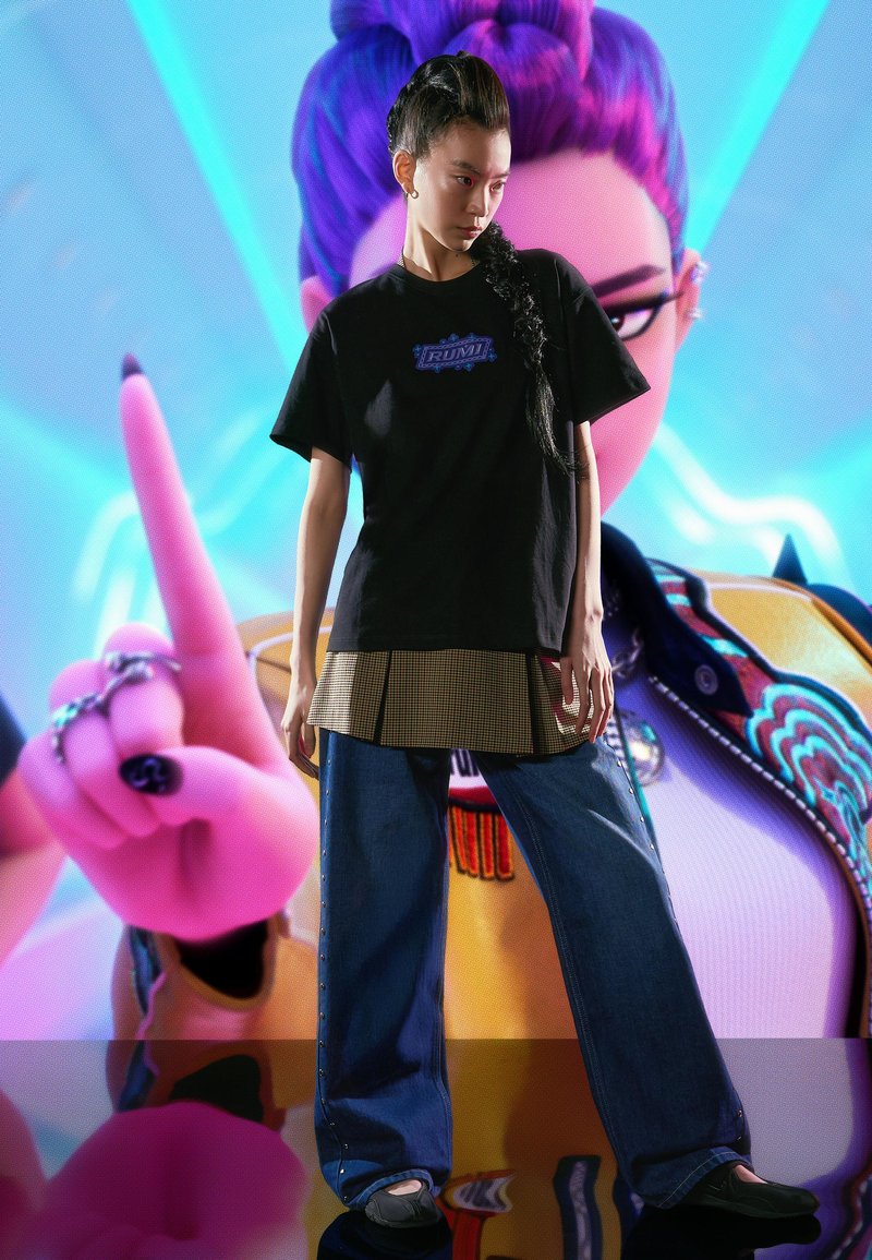 Fekete grafikás póló rétegezve egy kockás tunika fölött, széles szárú kék farmerrel párosítva. A modell fekete cipőt visel. Az élénk háttér stilizált karaktert ábrázol.