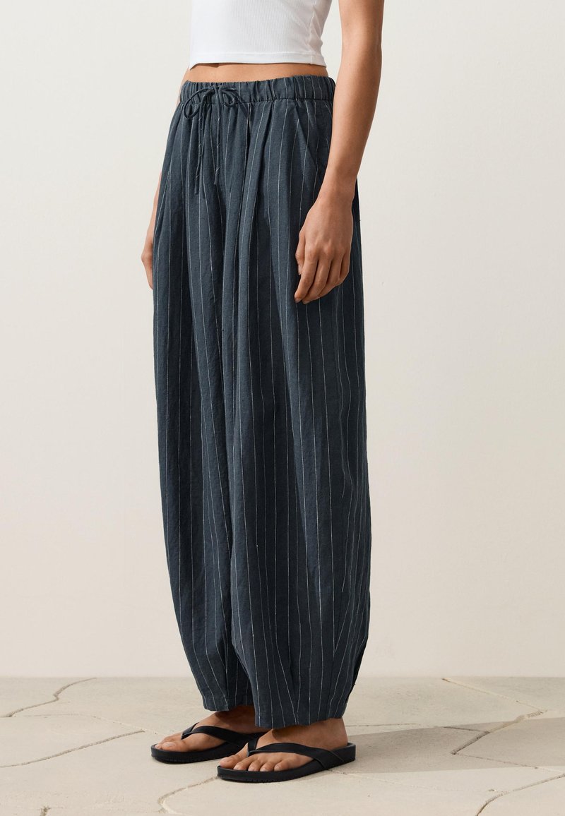 Personne portant un pantalon large bleu marine à fines rayures avec une taille élastique et des tongs noires sur un sol carrelé.
