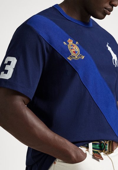 Μπλε ναυτικό κοντομάνικο πόλο με αντίθετη μπλε διαγώνια γραμμή, κεντημένες λεπτομέρειες και τον αριθμό «3» στο μανίκι.