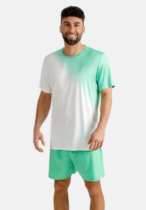 Lachende man met een groen-wit verlopend t-shirt en effen groene shorts, staand tegen een effen lichte achtergrond.