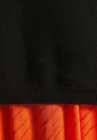 Haut noir en maille avec ourlet côtelé superposé à une jupe orange présentant des motifs ondulés diagonaux subtils, une texture lisse et un tissu léger.