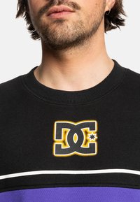 Černá mikina s výrazným logem DC v žluté a šedé barvě. Má texturovanou tkaninu a vodorovný bílý pruh nad fialovým pruhem.