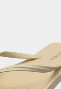 Tongs en caoutchouc beige avec semelle texturée et fines lanières, photographiées en gros plan sous un angle sur fond blanc.