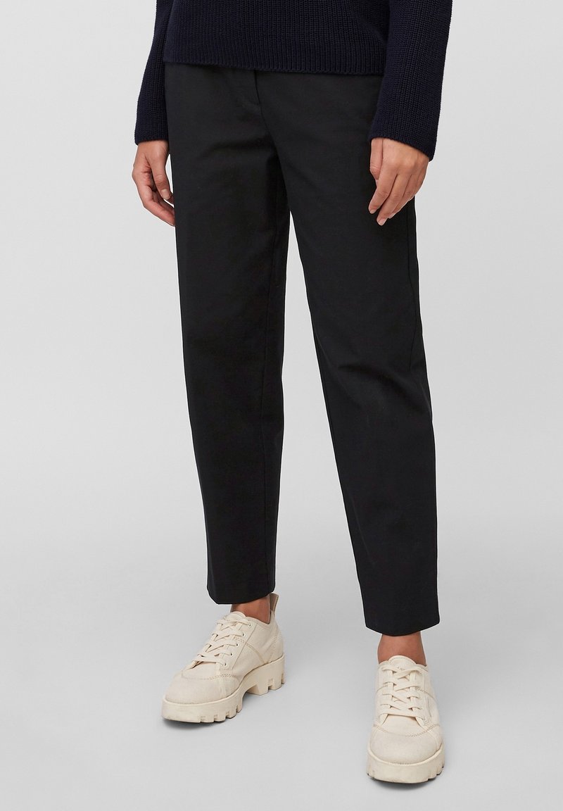 Persoon in zwarte tapse broek, off-white chunky sneakers en een marineblauwe trui die op een effen achtergrond staat.