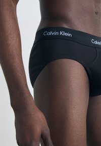 Schwarze Baumwollslips mit einem weichen Bund, der das "Calvin Klein"-Logo in Weiß trägt. Glatte Textur, figurbetontes Design und mittelhoher Schnitt.